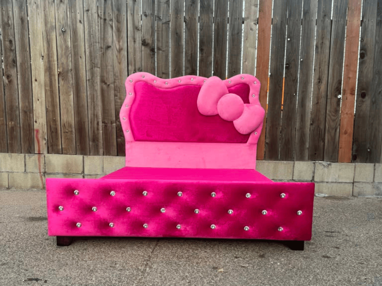 Hot pink velvet bed with a scalloped bow headboard and a crystal-button tufted footboard, photographed outdoors against a timber fence