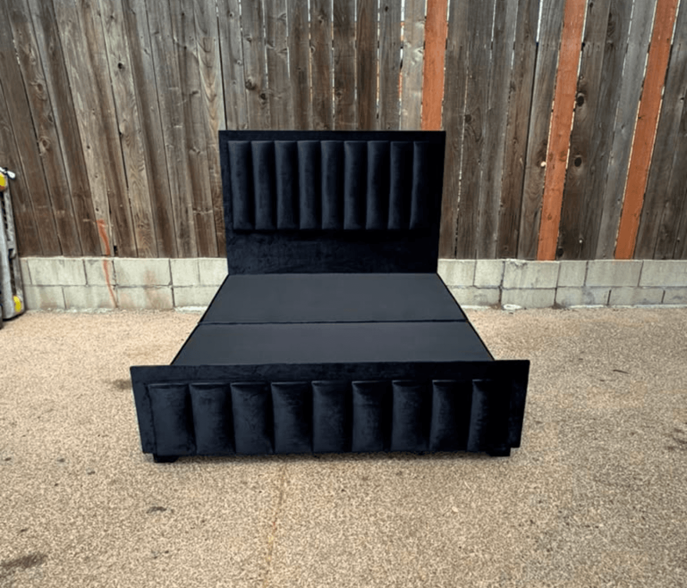 Black velvet bed with a vertical channel-tufted headboard and a matching panelled footboard, against a timber fence