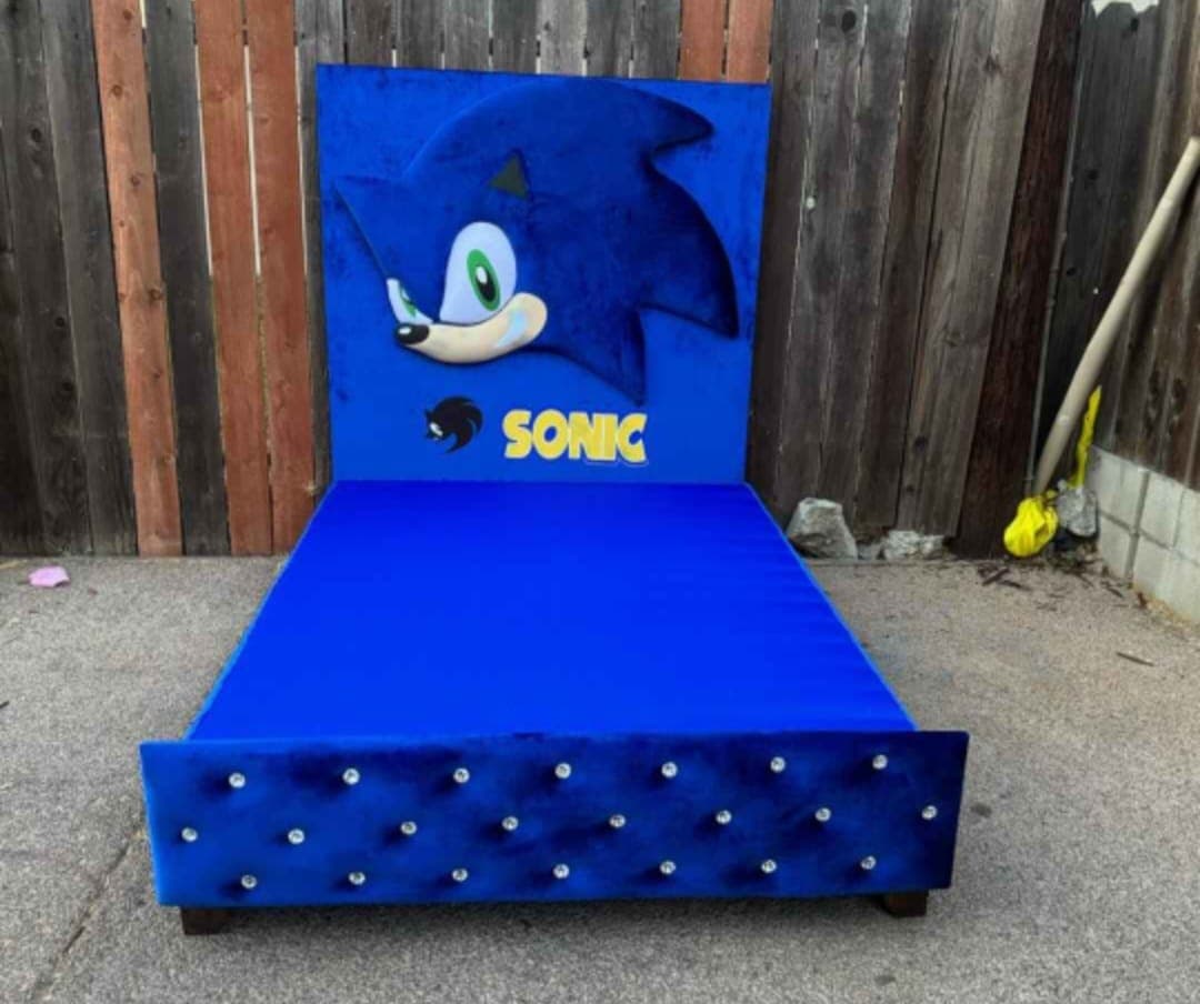 Sonic Bed head-on, a tall blue velvet headboard carrying a sculpted blue character face above a matching base and a crystal-buttoned footboard